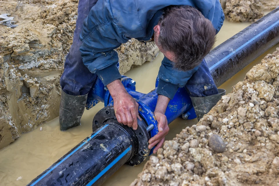 Ouvrier en combinaison bleue installant une canalisation d'eau dans une tranchée boueuse