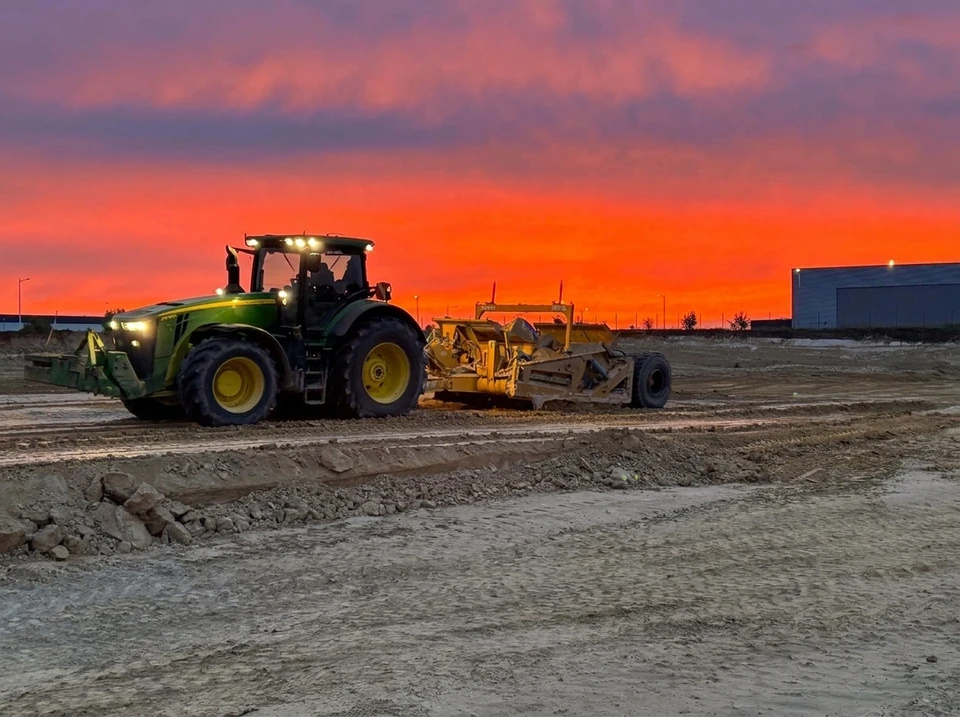 Photographie d'un scraper tracté en action sur un chantier de terrassement, montrant la machine décapant et nivelant la terre sur une grande surface. La machine doit être clairement visible avec son système de traction et de décapage en fonctionnement. Le chantier occupe l'arrière-plan avec de larges surfaces de terre remaniée, montrant les volumes importants déplacés. Lumière naturelle de jour, image nette et professionnelle.