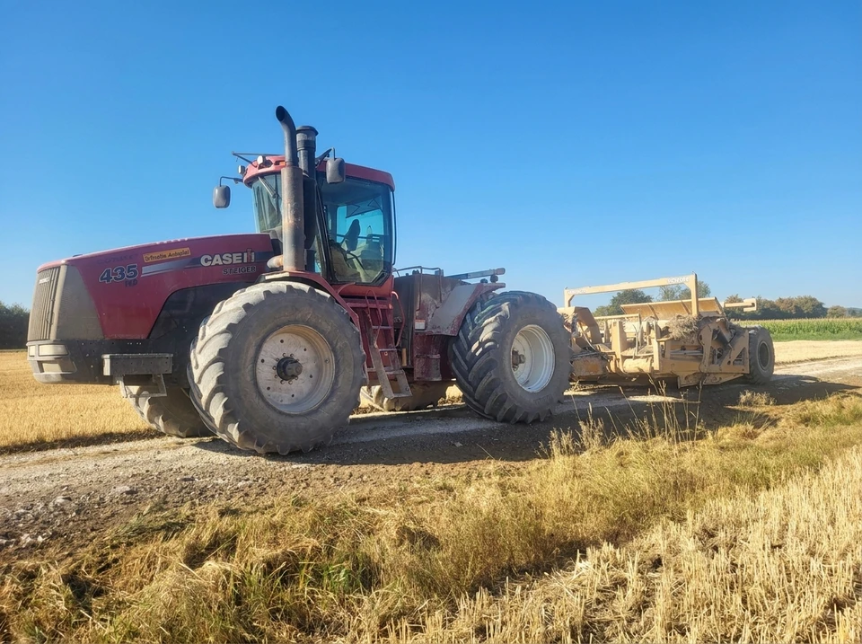 Photographie réaliste d'un scraper tracté en action sur un chantier de terrassement professionnel. La machine, impressionnante par sa taille et sa structure, travaille sur un terrain dégagé, effectuant un décapage de sol. L'engin est clairement visible de profil ou légèrement de trois-quarts, montrant ses caractéristiques distinctives. L'arrière-plan montre un chantier actif avec un ciel dégagé. La photo est prise en lumière naturelle de jour, avec une qualité professionnelle nette et détaillée.