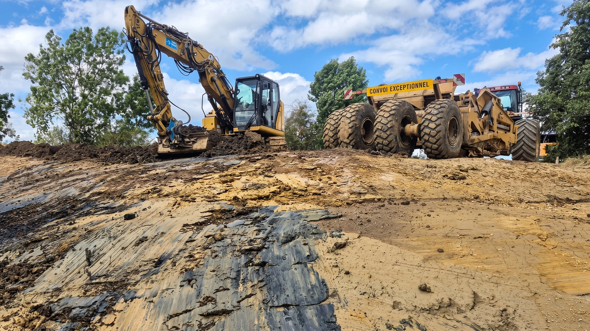 Chantier de terrassement de grande envergure en extérieur photographié de jour, montrant un scraper tracté (engin articulé jaune et noir de grande dimension) en action sur un vaste terrain, soulevant et transportant de la terre, avec un ciel dégagé et du terrain excavé visible en arrière-plan. L'image doit clairement mettre en avant la machine distinctive et sa capacité de travail impressionnante.